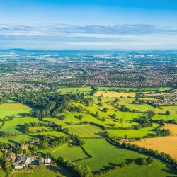 Aerial view over picturesque patchwork landscape of green pasture, lush meadows and crop fields, farmhouses and rural homes amongst the rolling hills and quiet valleys below panoramic blue summer skies. ProPhoto RGB profile for maximum color fidelity and gamut.